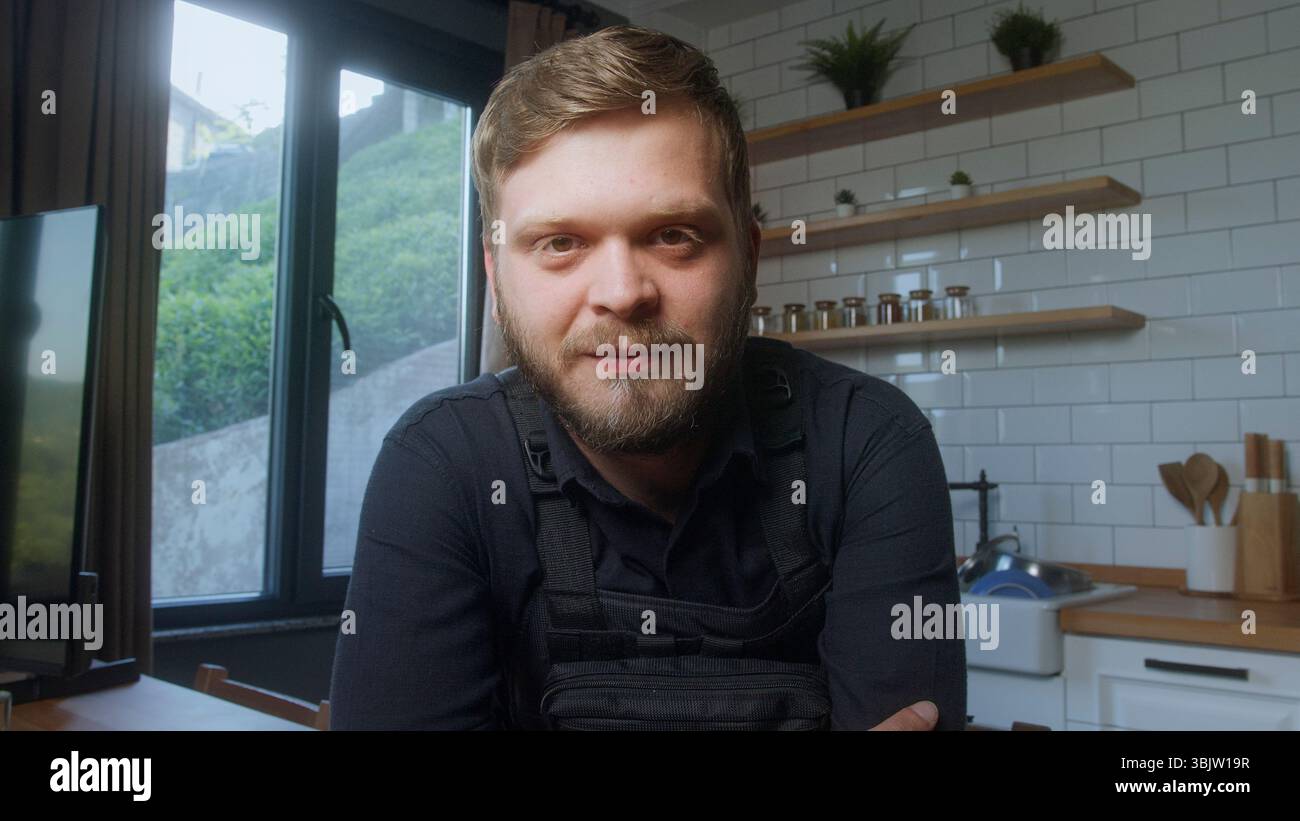 Bearded man wearing a black tactical vest looks directly at the camera with a calm, confident expression in a modern indoor setting. Stock Photo