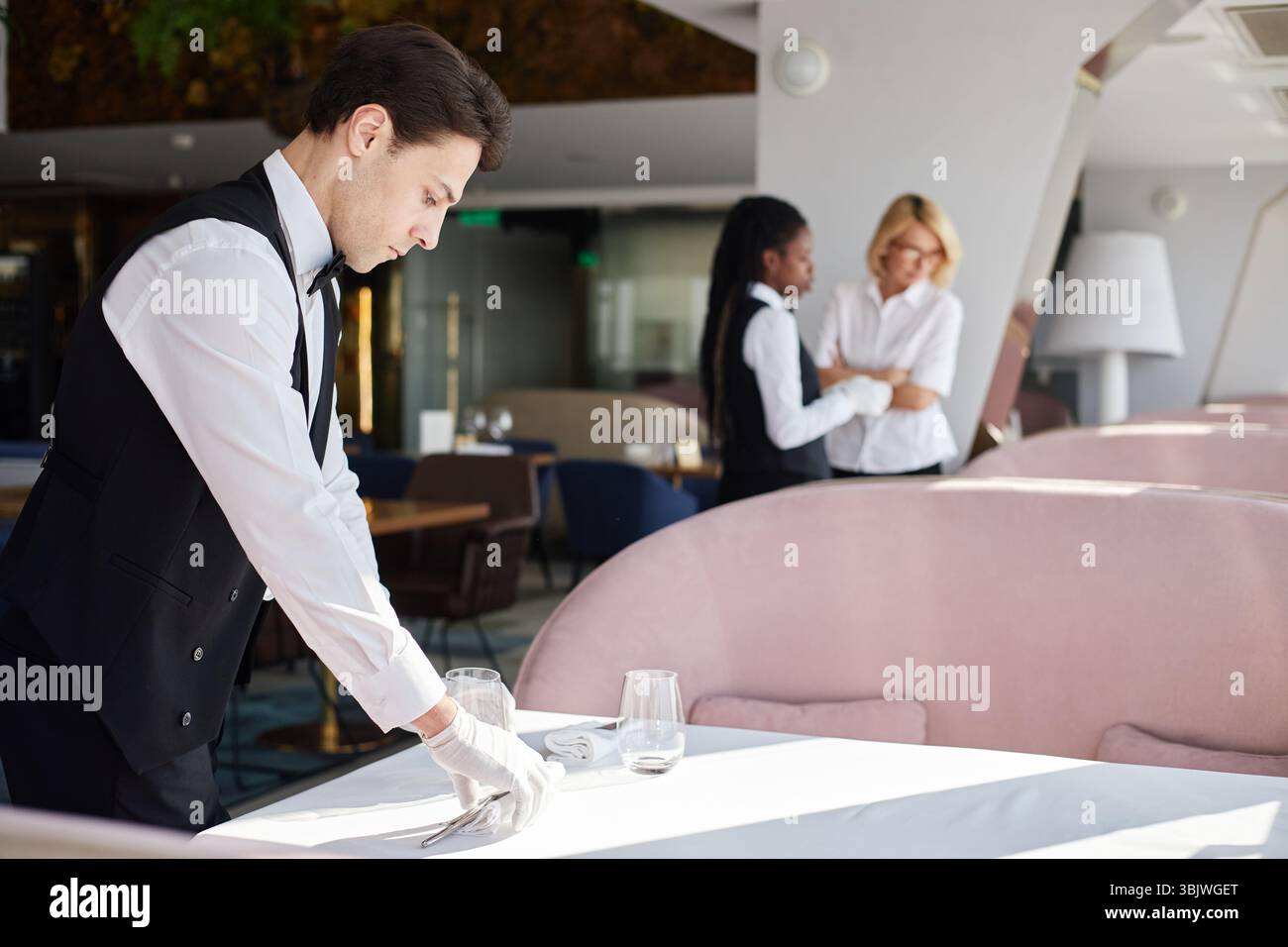 Young Caucasian Man Setting Table While Two Female Coworkers Discussing Work Stock Photo