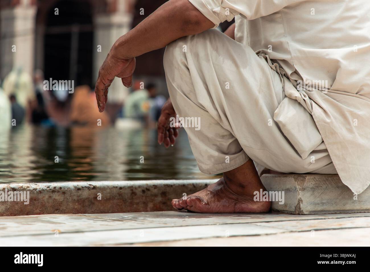 Barefoot indian man wearing traditional clothes resting by the water in Jama Masjid Mosque, the main mosque of Old Delhi, India, during a sunny day, w Stock Photo