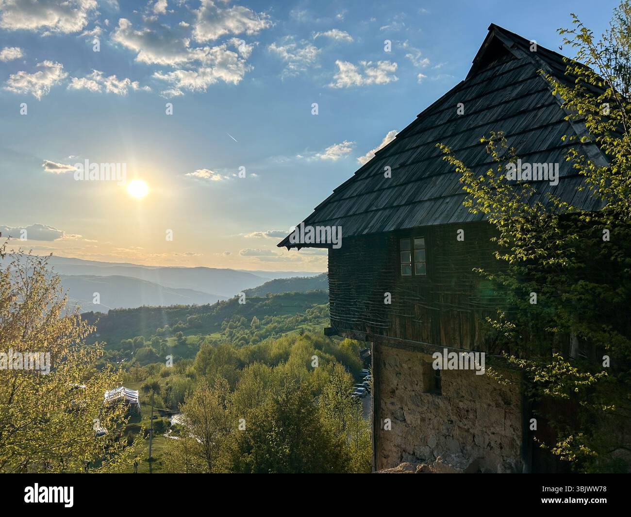 Scenic view of a traditional wooden mountain house at sunset, overlooking a lush green valley and distant hills under a glowing sky. - Smartphone Captured Stock Image