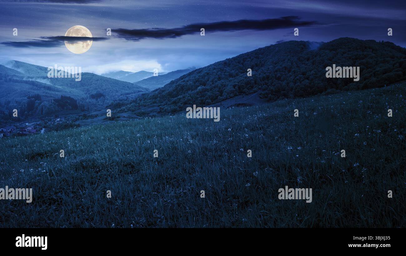 rural landscape in mountain at night. traditional pasture on the grassy slope in full moon light. starry sky with dark clouds Stock Photo