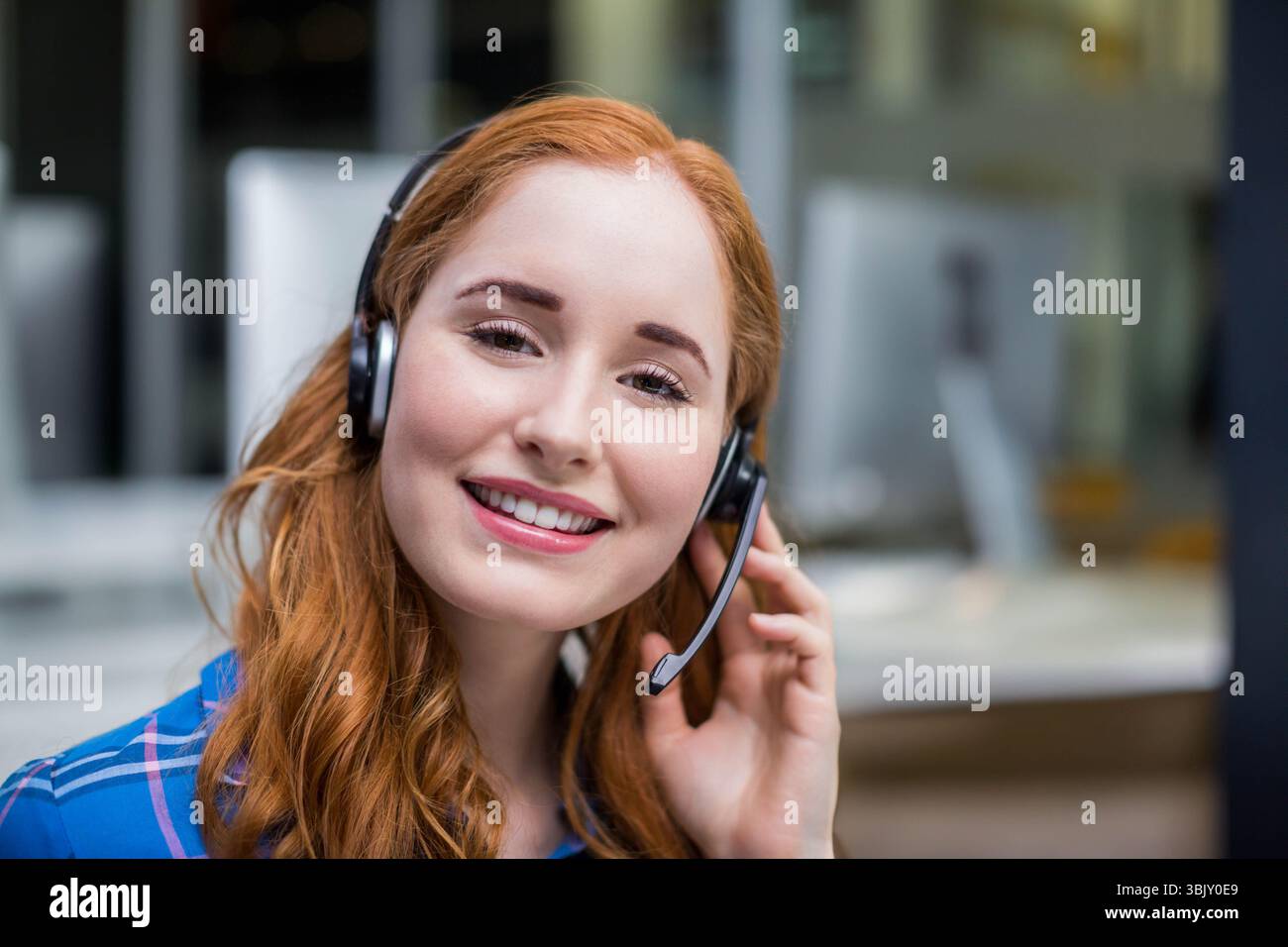 Red-haired support rep using headset and blue plaid shirt smiling at open-plan office environment Stock Photo