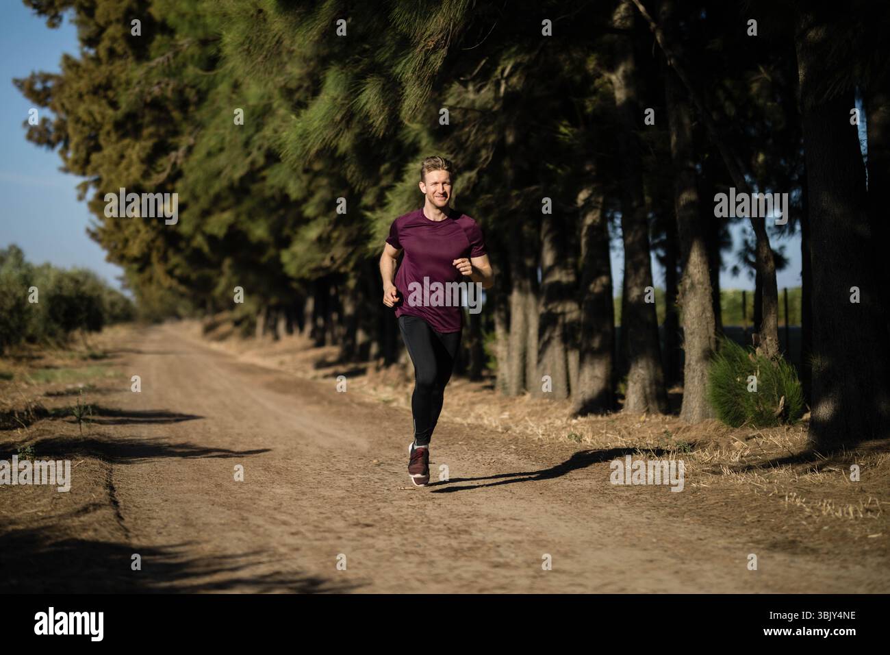 Male jogger running along dirt trail through pine forest wearing maroon athletic shirt Stock Photo
