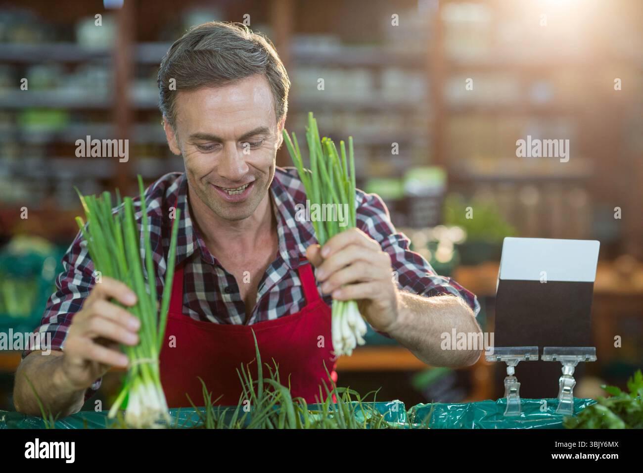 Male vendor arranging green onion bunches at market stall wearing red ...
