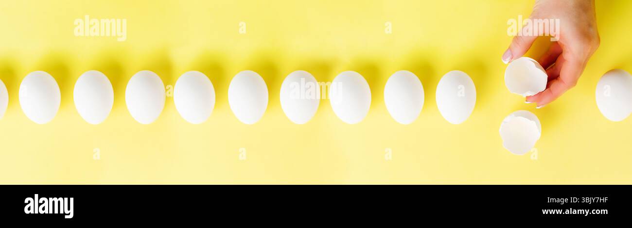 White raw chicken eggs lying in row on a yellow trendy background and female hand holds broken egg. Easter minimal creative comp Stock Photo