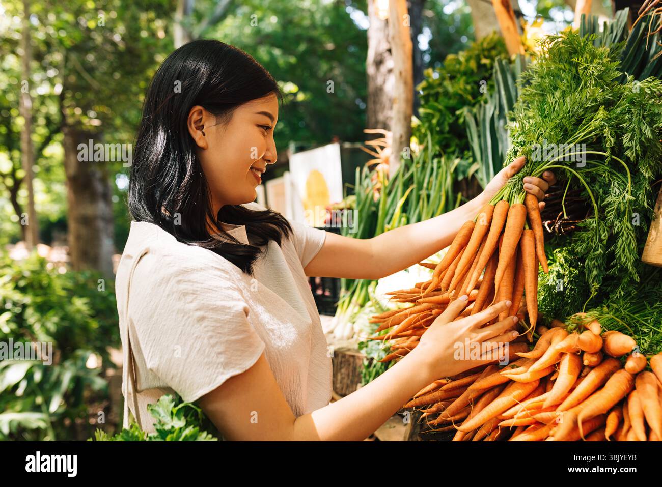Smiling buyer holding a bunch of carrots. Asian woman choosing vegetables in the local market. Stock Photo