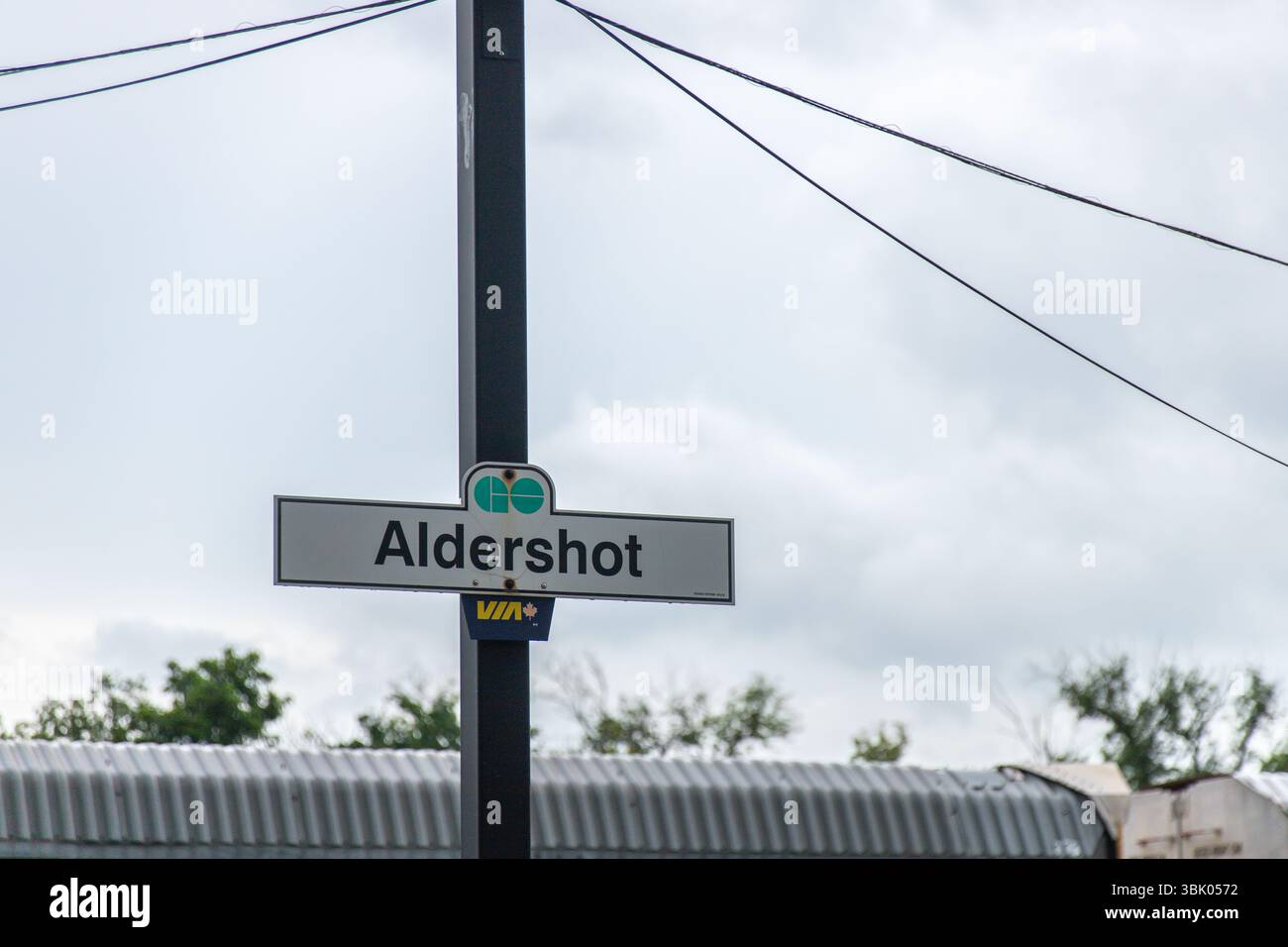 Aldershot train station sign featuring GO Transit and VIA Rail logos, set against a cloudy sky and industrial train yard background. Stock Photo