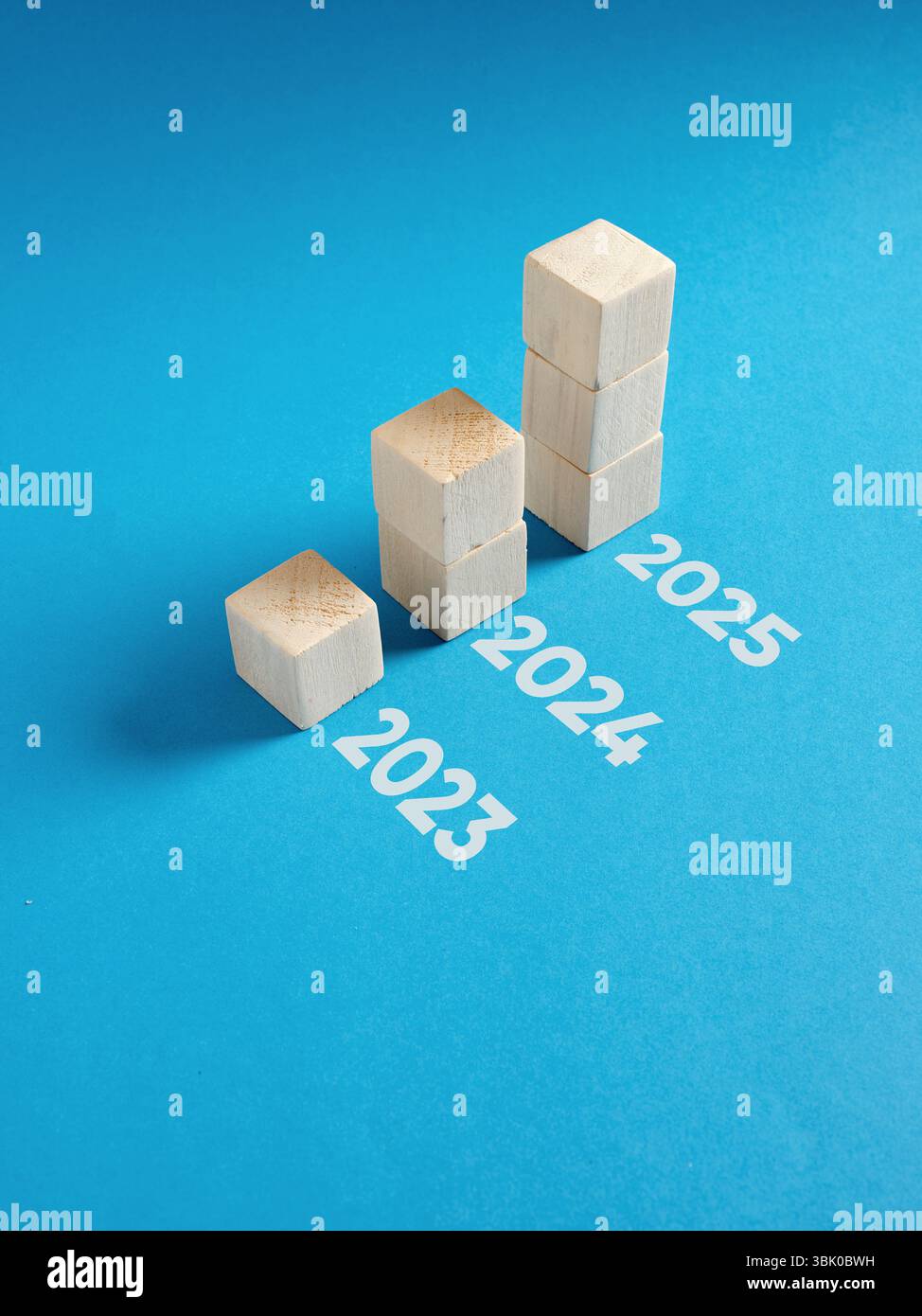 Concept of business growth over time with wooden cubes forming an ascending bar chart from year 2023 to 2025 on blue background Stock Photo