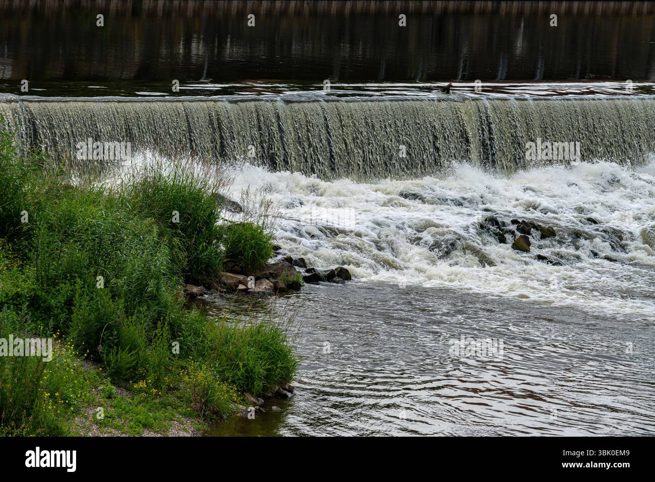 Water rushes over a dam, creating white frothy waves at the base ...