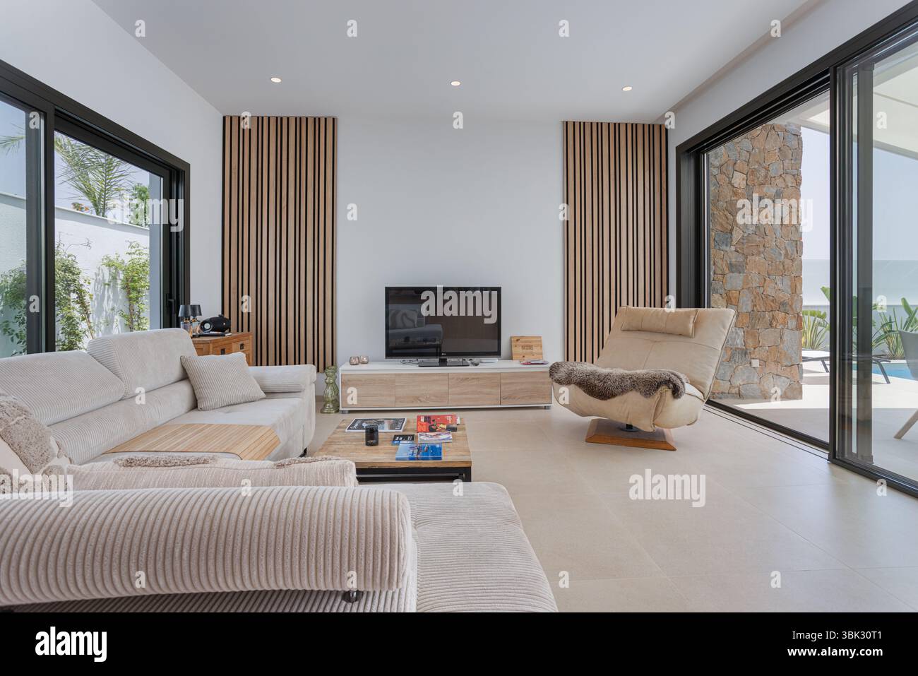 Bright and inviting living space with TV, stylish panel accents, and sliding doors opening to a poolside terrace with lounge furniture. Stock Photo