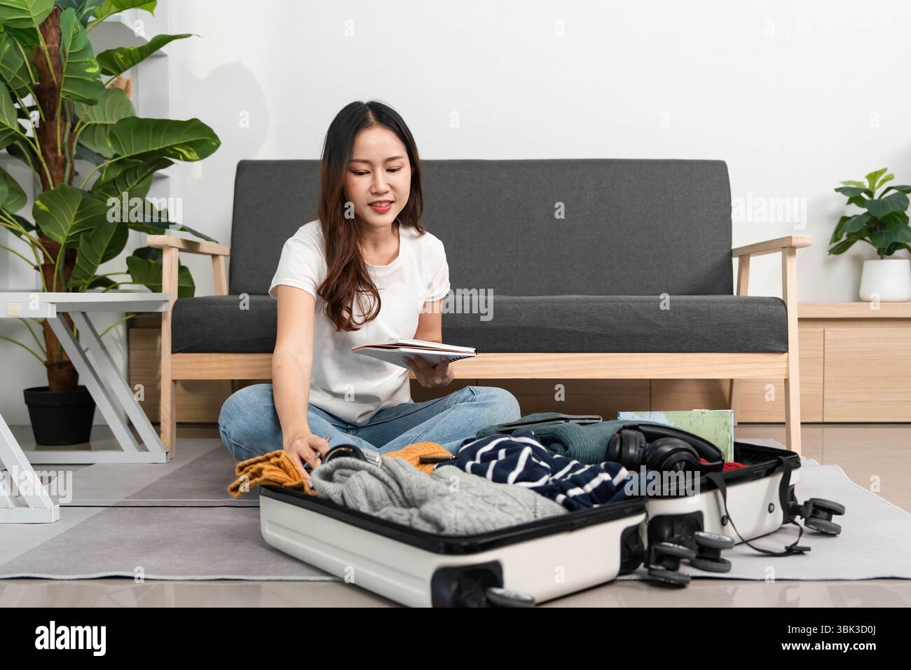 Travel Preparation. A woman sits on the floor, reviewing her packing list while organizing her suitcase. Stock Photo