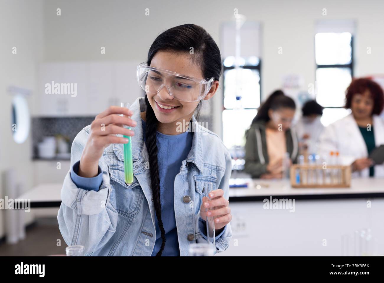 Girl in science class examining test tube, smiling and wearing safety goggles, at school Stock Photo