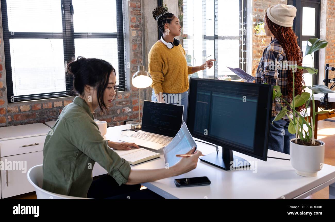 Collaborating on software, diverse women using laptops and monitors in modern office Stock Photo