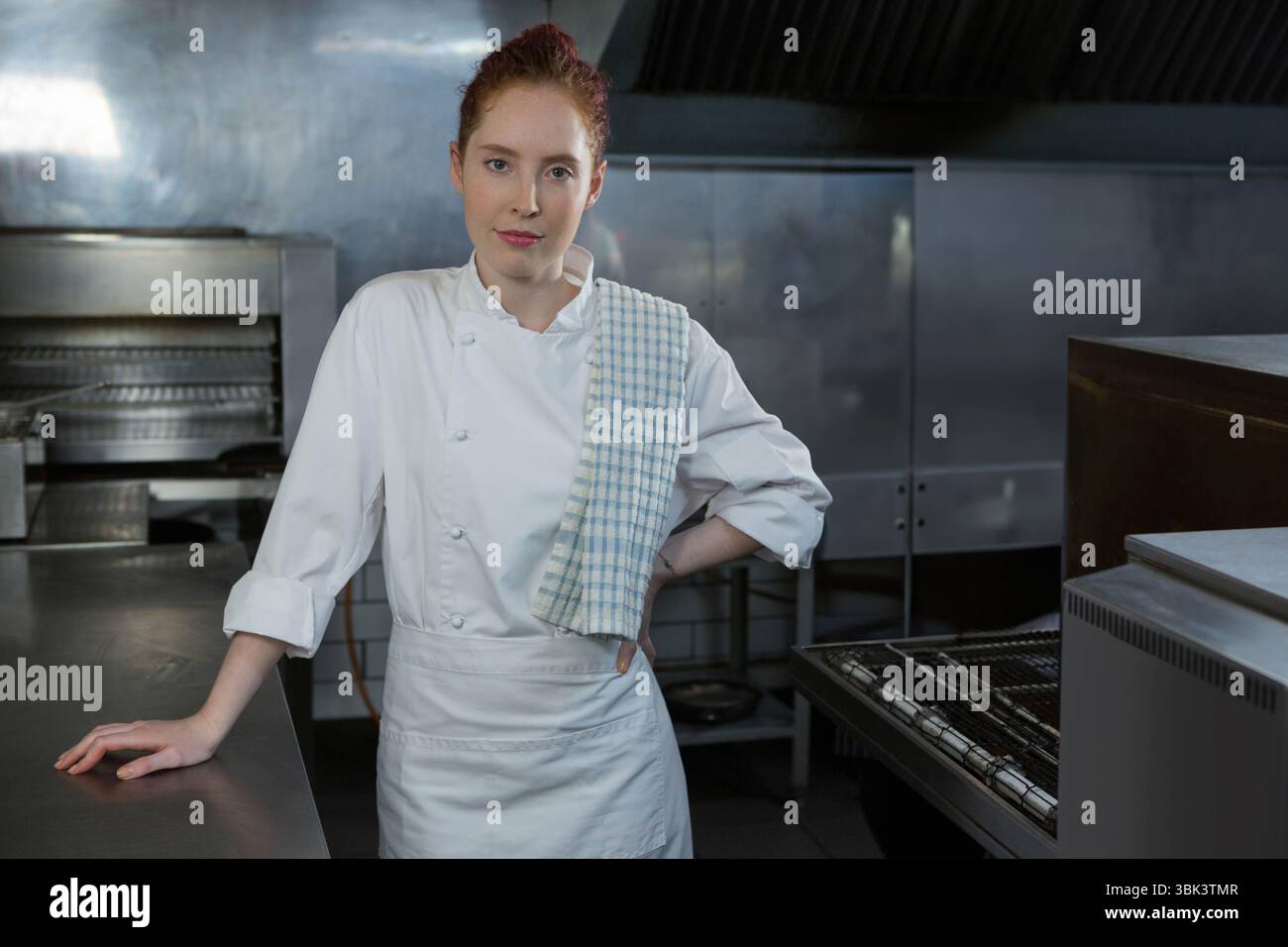 Female chef wearing chef jacket and checkered towel leaning on counter in kitchen with ovens Stock Photo
