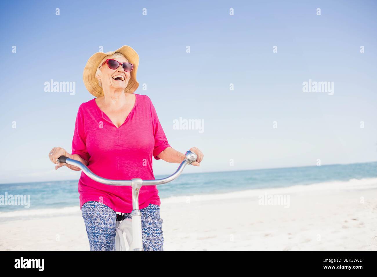 Senior woman riding cruiser bicycle laughing along sandy beach wearing ...