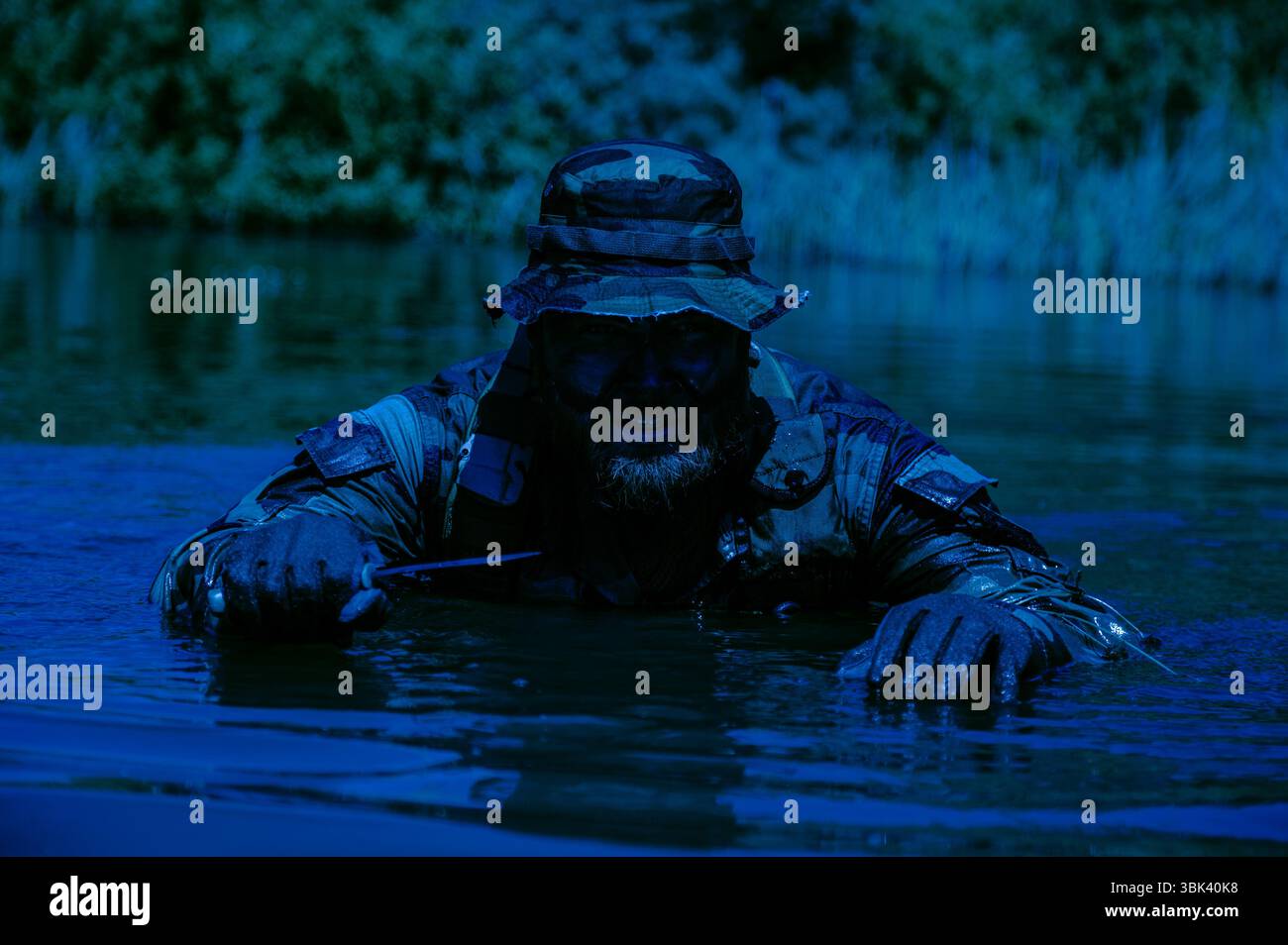 Man in camouflage clothing and hat crouching in water holding a knife ...