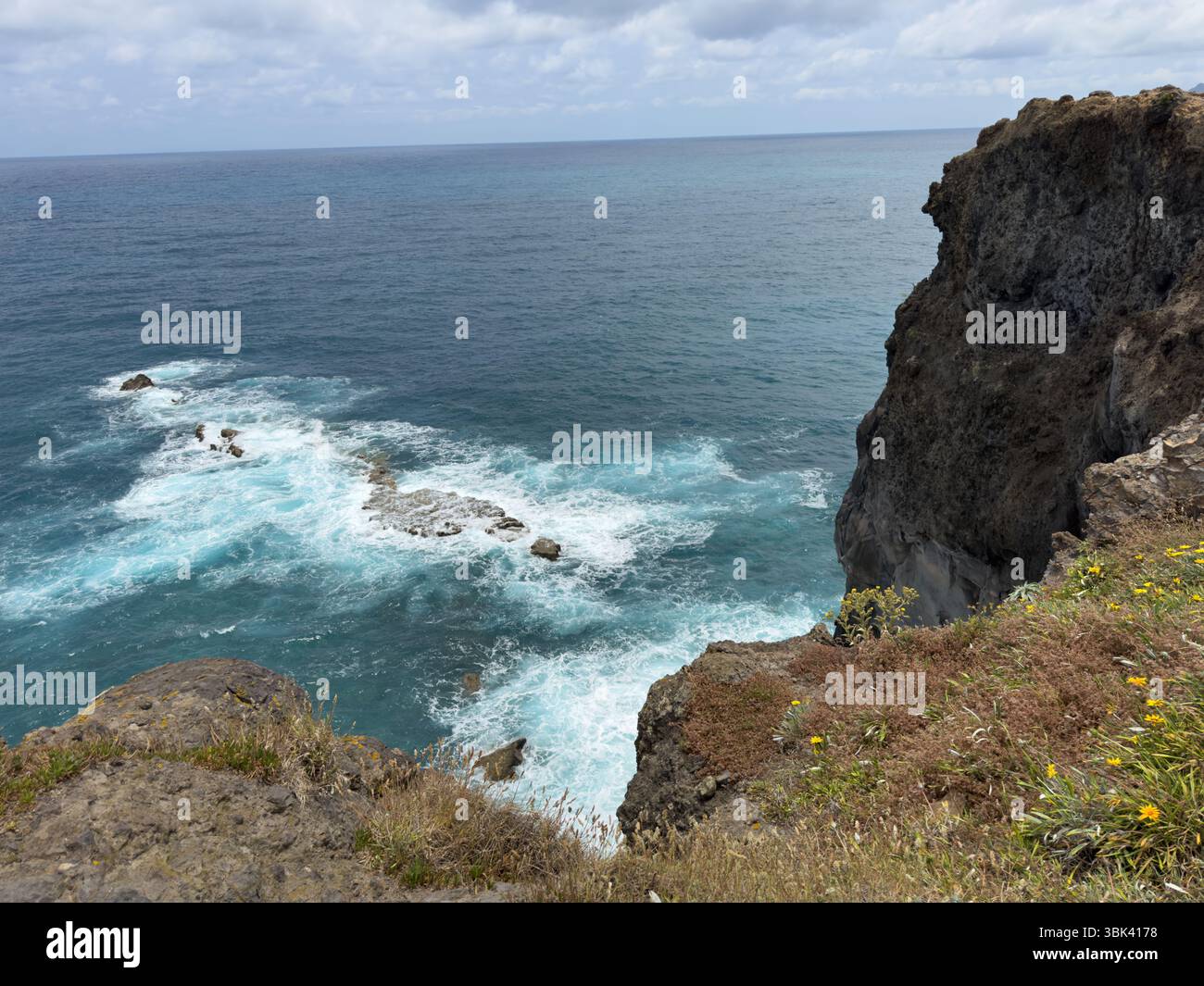 Ocean waves crash against the rocky coastline of Madeira, Portugal. The seascape features white surf, sea spray, and rugged stones. - Smartphone Captured Stock Image
