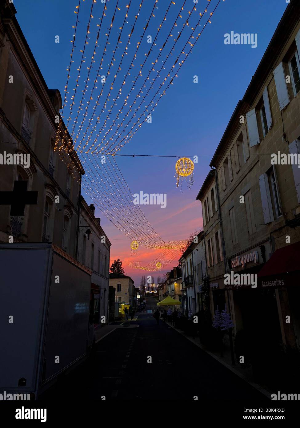 Charming French village street at twilight with festive Christmas lights and a vibrant sunset sky in the background - Smartphone Captured Stock Image