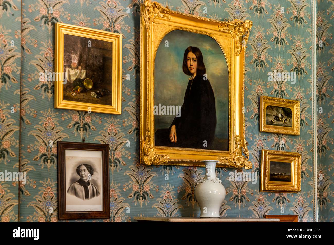 The salon in the George Sand House (Maison de George Sand) on Place Sainte-Anne in Nohant, municipality of Nohant-Vic, in the Berry, Indre department, Centre-Val de Loire, France, displays portraits of George Sand and her family, with a large portrait of George Sand as a young woman in the centre. The painting in an ornate gilded frame shows her seated in a black dress with dark, chin-length hair. A still life hangs on the left, a small monochrome portrait of the older writer below it, and two small landscapes on the right. A white vase stands on the mantelpiece in front of blue floral wallpap Stock Photo