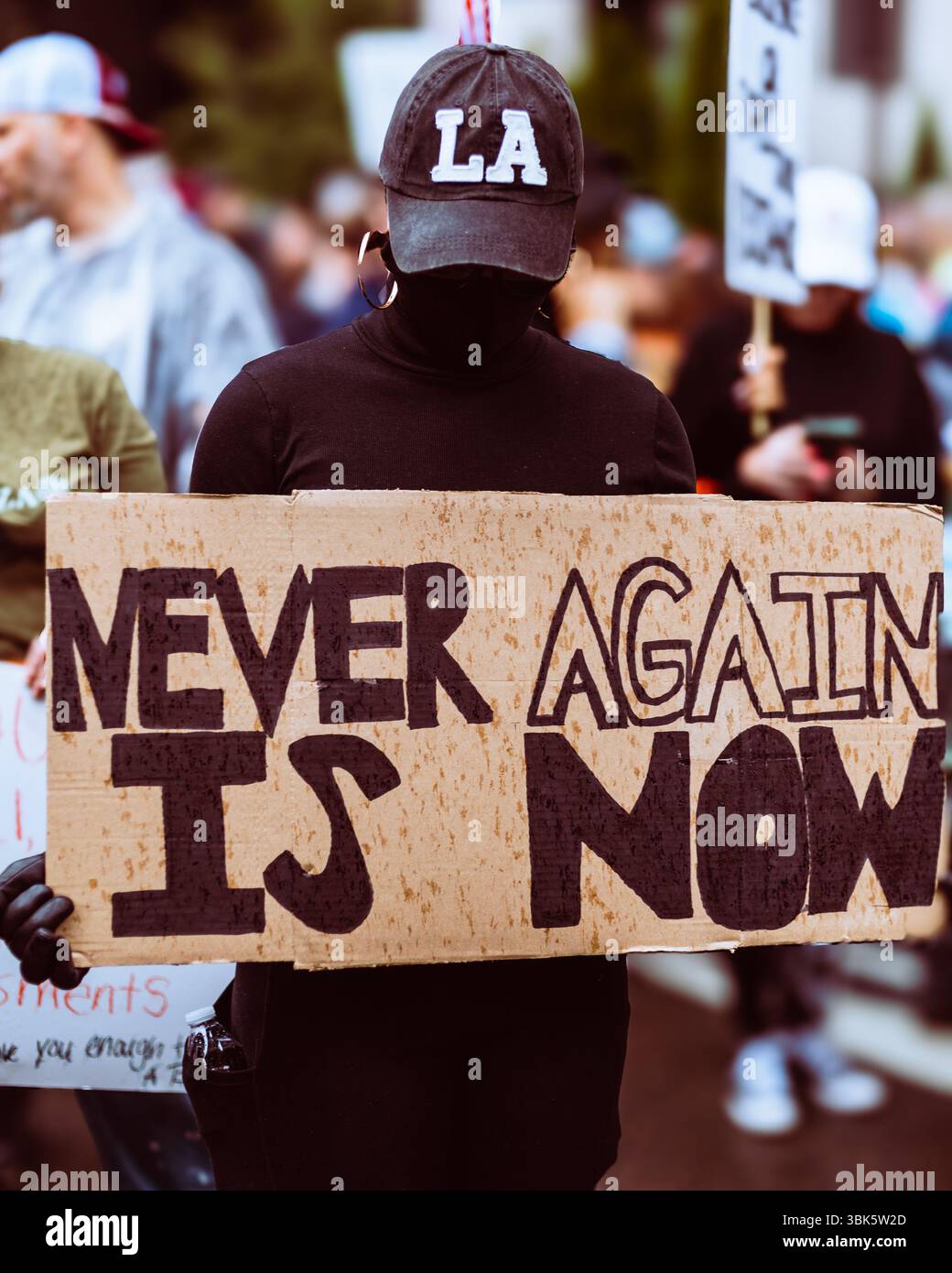Woman wearing LA hat holds "Never Again is Now" sign during "No Kings ...