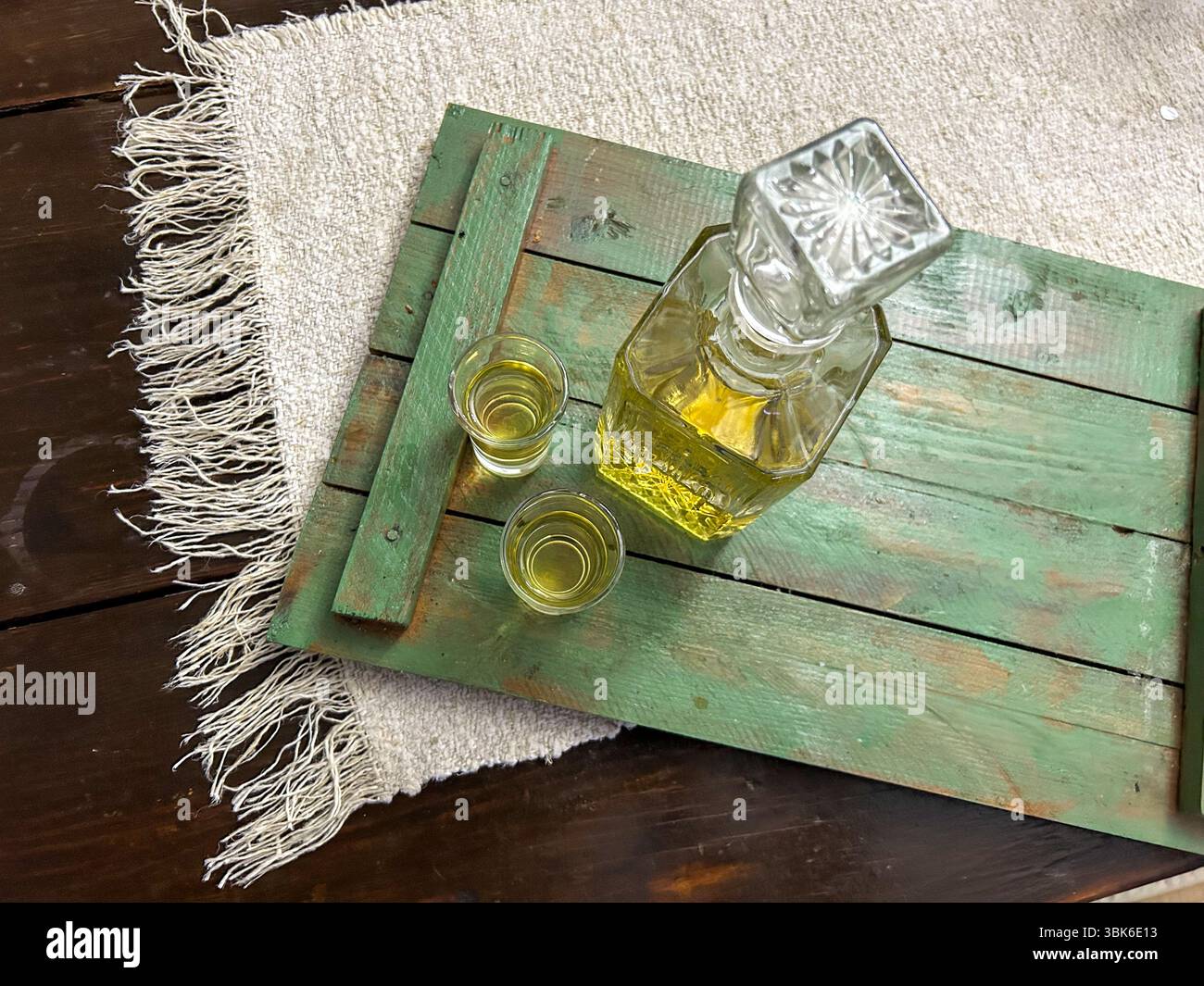 A decanter and two shot glasses filled with homemade rakija on a vintage green wooden tray and woven cloth, reflecting Balkan tradition and hospitalit - Smartphone Captured Stock Image