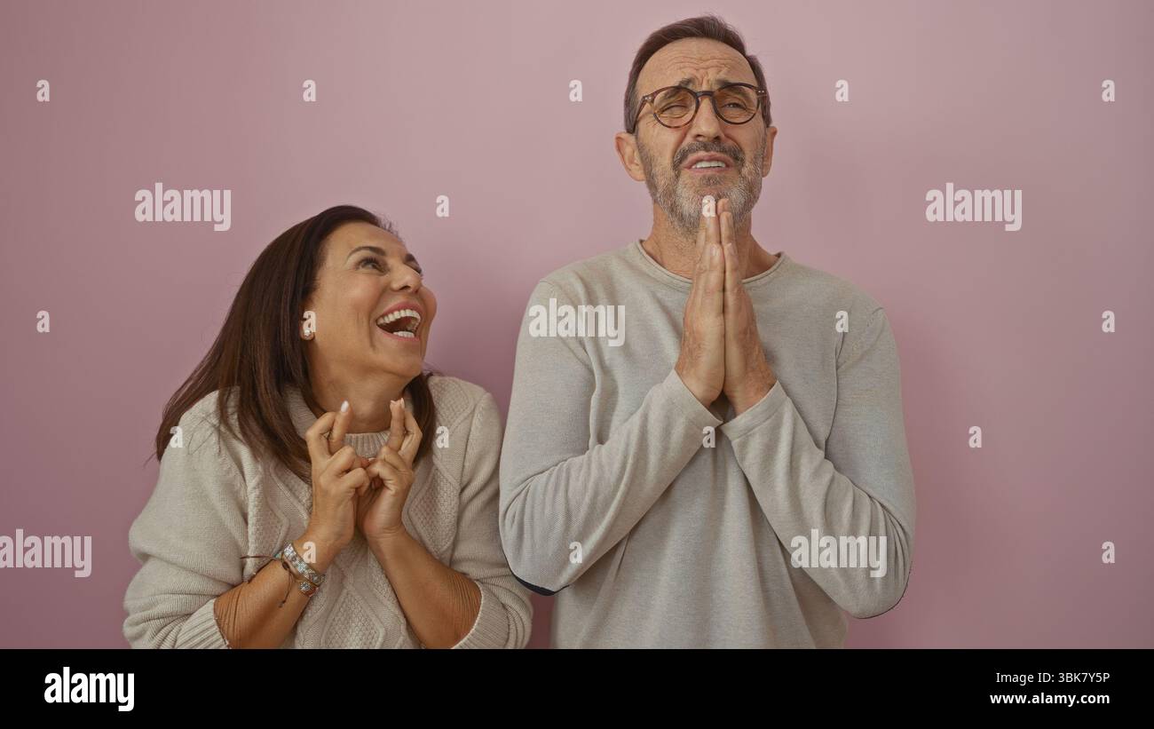 Middle-aged hispanic couple expressing love and humor with fingers crossed and hands together against a pink wall. Stock Photo