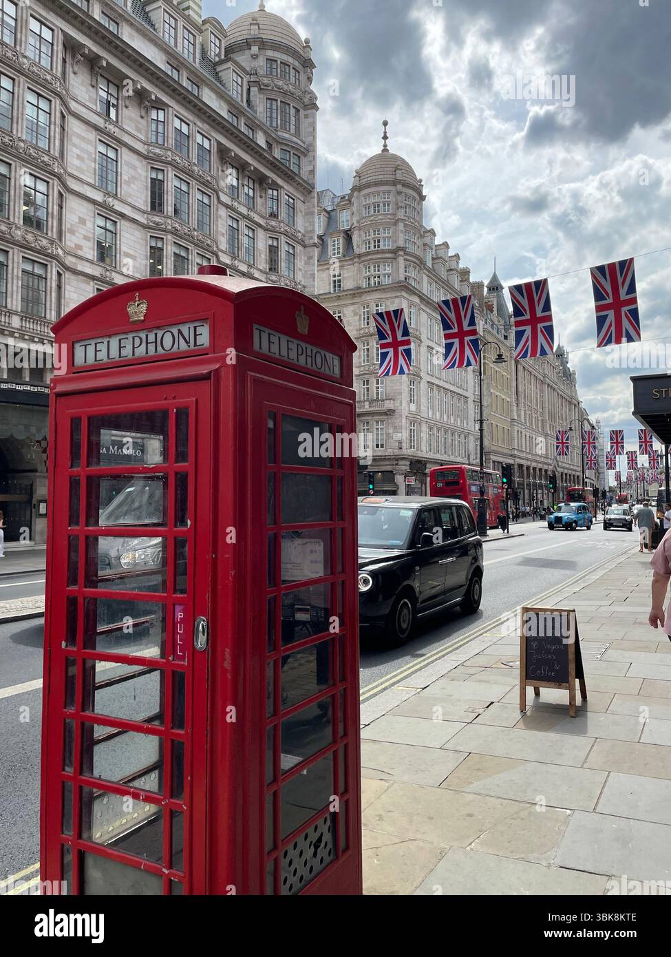 Iconic red telephone booth on a central London street with Union Jack flags and historic buildings in the background - Smartphone Captured Stock Image