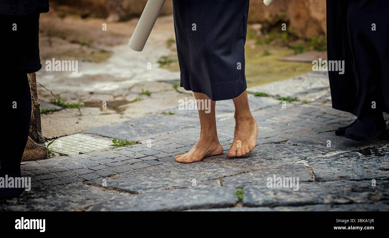 Barefoot Member Of A Religious Order Walking On Cobblestones During A Good Friday Procession, Carrying A Rolled Up White Cloth And Embodying Deep Fait Stock Photo