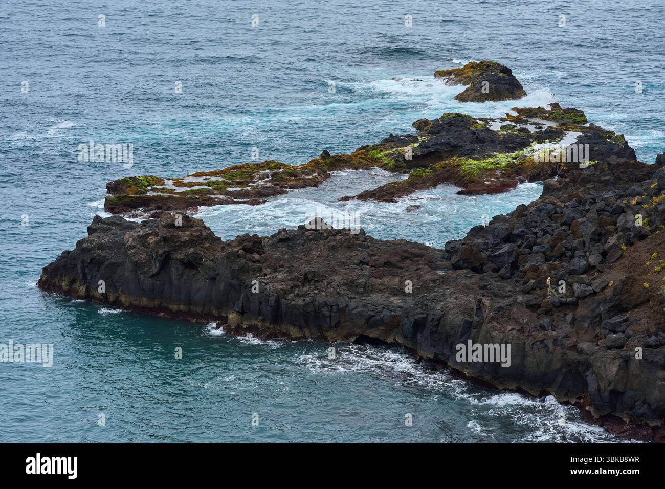 Aerial view of volcanic rock formations and ocean waves near El Bollullo beach in Tenerife, with foamy surf crashing against dark boulders and mossy s Stock Photo