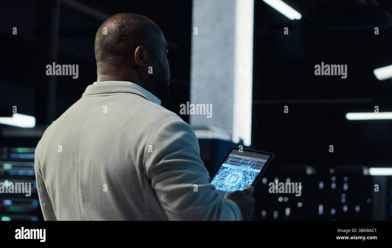 Server room software developer troubleshooting errors using software on laptop. African american IT professional walking in data center fixing bugs using notebook, camera B Stock Photo