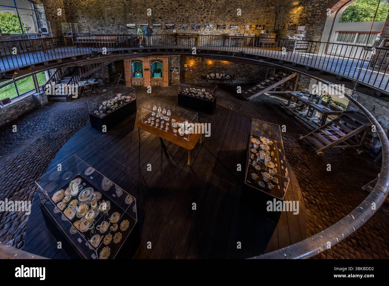 A temporary exhibition of Art Deco porcelain fills a round room in the Four des Casseaux museum (Musée du Four des Casseaux) on Rue Donzelot in Limoges, Haute-Vienne, Nouvelle-Aquitaine, France. Cups, plates and pots stand in glass display cases on a dark wooden floor, with a gallery with an iron railing running above. On the gallery, two visitors look at pictures on the rubble-stone wall. The exhibition is possibly "Limoges Art Déco 1925/2025", with around 200 porcelain pieces marking the centenary of the 1925 International Exhibition of Modern Decorative and Industrial Arts in Paris. The mus Stock Photo