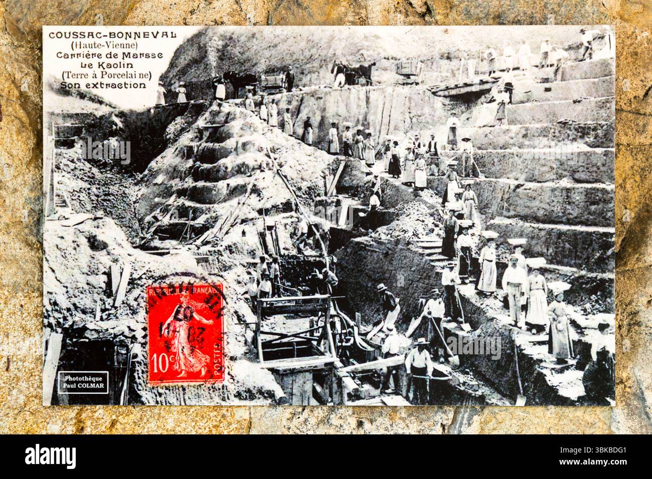 An old postcard with a motif from Coussac-Bonneval shows the extraction of kaolin. It hangs in the Musée du Four des Casseaux on Rue Donzelot in Limoges, Haute-Vienne, Nouvelle-Aquitaine, France. The caption reads "Coussac-Bonneval (Haute-Vienne) – Carrière de Marsse – Le Kaolin (Terre à Porcelaine) – Son extraction". In English: "Marsse quarry – kaolin (porcelain clay) – its extraction". Men in hats and women in long skirts work on the stepped terraces of the pit, and wooden scaffolding stands in the foreground. At the top edge, horses stand next to a cart. A red 10-centime stamp is stuck on Stock Photo