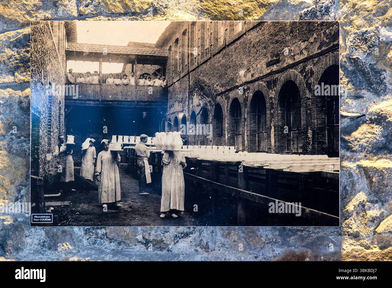 An old postcard in the Four des Casseaux museum (Musée du Four des Casseaux) on Rue Donzelot in Limoges, Haute-Vienne, Nouvelle-Aquitaine, France, shows a photograph by Paul Colmar: female workers at a porcelain factory transporting blanks. Women in long, light-coloured smocks and headscarves carry tall stacks of white blanks on their heads and shoulders across a factory yard. A man in an apron stands among them. On the right, long wooden racks with rows of white pieces stand in front of a brick building with arched windows. Workers look down from a wooden gallery above. The note "Photothèque Stock Photo