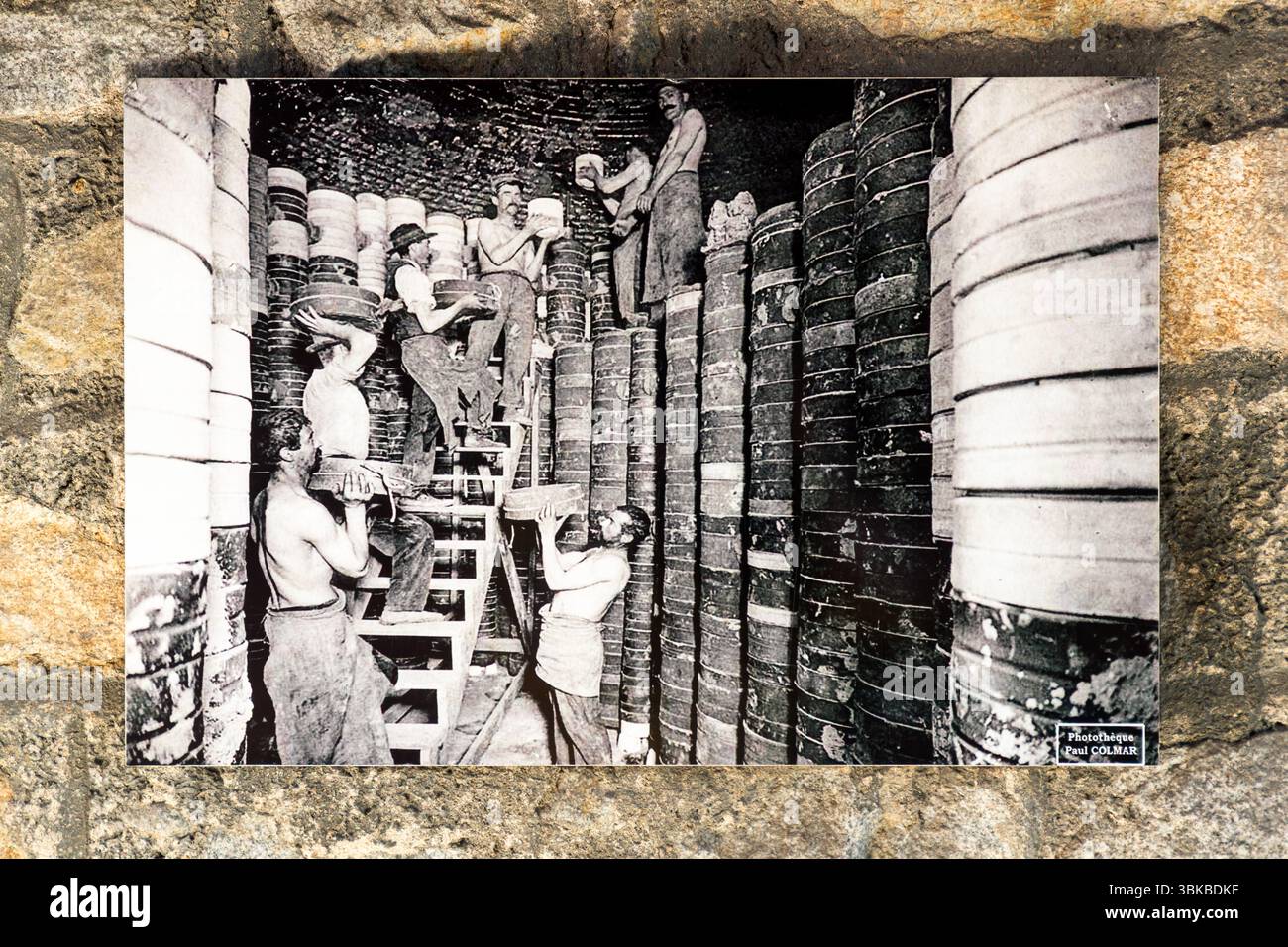An old postcard in the Four des Casseaux museum (Musée du Four des Casseaux) on Rue Donzelot in Limoges, Haute-Vienne, Nouvelle-Aquitaine, France, shows a photograph by Paul Colmar: factory workers packing the kiln for the second firing. The heavy clay moulds (saggers, French gazettes), each containing a porcelain blank, were stacked metres high in the large kiln. Men, some of them bare-chested, lift the saggers upwards via a wooden ladder. Columns of saggers reach up to the brick vault of the kiln. The note "Photothèque Paul Colmar" appears at the bottom right. Depending on the type of porcel Stock Photo