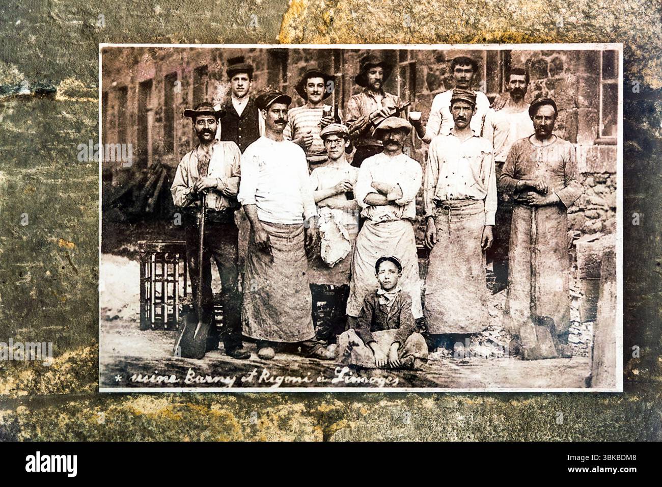 A group picture of workers at the kiln of a porcelain factory in Limoges, Haute-Vienne, Nouvelle-Aquitaine, France, hangs in the Four des Casseaux museum (Musée du Four des Casseaux) on Rue Donzelot. The workers' postures and facial expressions are unusually relaxed and individual. The photographer is unknown. The handwritten caption reads "usine Barny et Rigoni à Limoges". In English: "Barny and Rigoni factory in Limoges". Men in long aprons, shirts and hats stand in front of a stone building, two of them holding shovels. A boy sits on the ground at the front. The museum preserves a porcelain Stock Photo