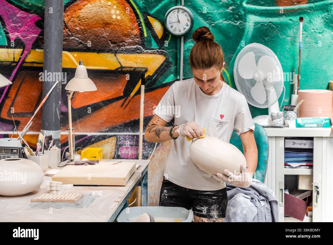 At the Ateliers Arquié art porcelain workshop on Avenue Baudin in Limoges, Haute-Vienne, Nouvelle-Aquitaine, France, a craftswoman smooths a large, egg-shaped porcelain body with a sponge. She wears disposable gloves and a white T-shirt with a small red heart, and her forearm is tattooed. A wall clock and a fan hang behind her in front of graffiti in green, orange and purple. The workshop was founded in 1996 and specialises in the design and production of customisable and artistically designed porcelain objects. It combines traditional craftsmanship with modern shapes, colours and techniques. Stock Photo