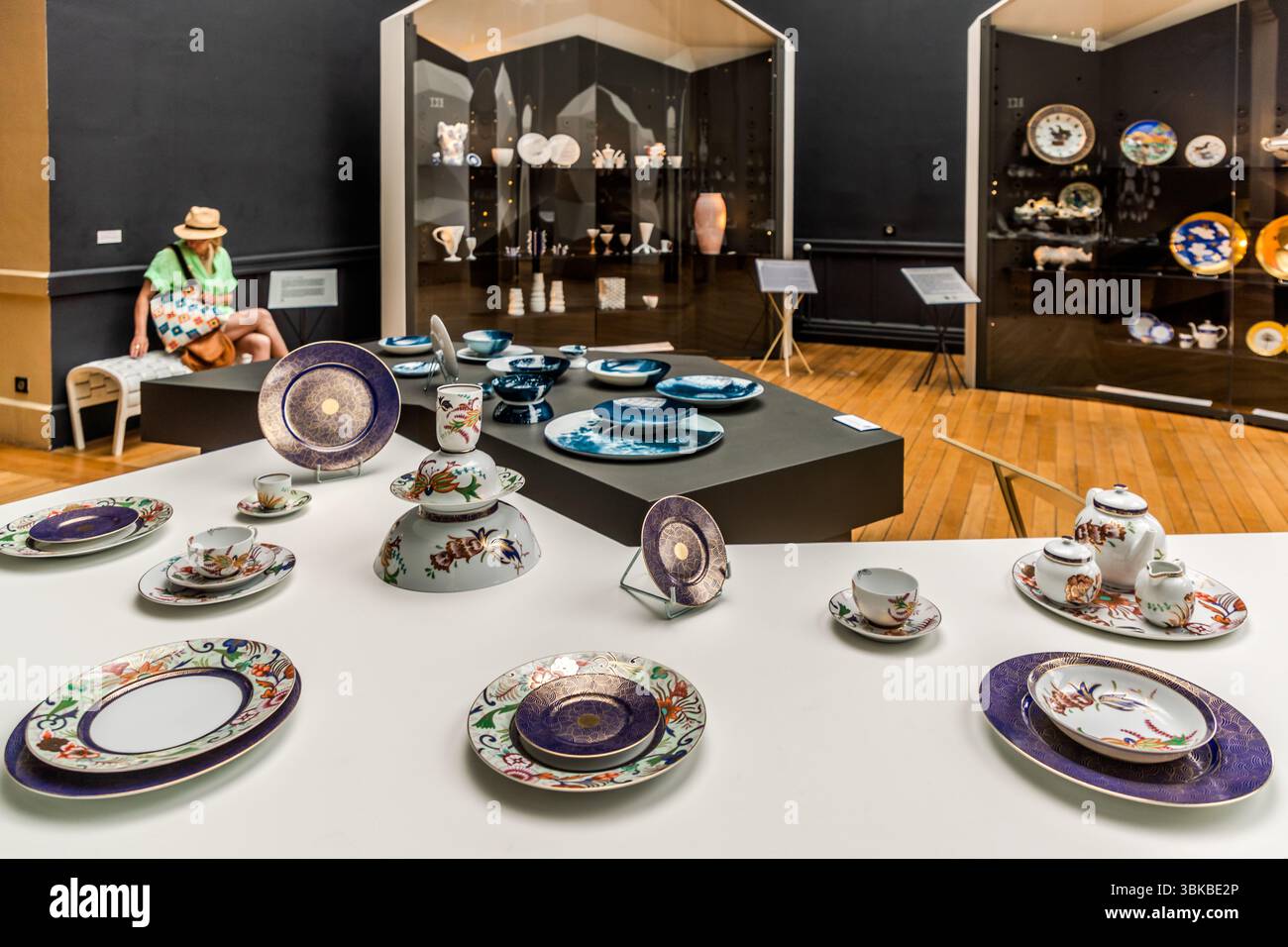An exhibition in the Musée National Adrien Dubouché on Place Winston Churchill in Limoges, Haute-Vienne, Nouvelle-Aquitaine, France, shows porcelain series from well-known brands such as Bernardaud (formerly Manufacture Royale de Limoges), Haviland, Jacques Pergay, Raynaud and Ateliers Arquié. Table settings stand on a white table: plates with purple and gold rims, plates and cups with colourful floral motifs, and a teapot with a milk jug and sugar bowl. Behind them, blue plates lie on a dark platform, and the display cases hold wall plates, figures and vases. Journalist Angela Berg sits on a Stock Photo