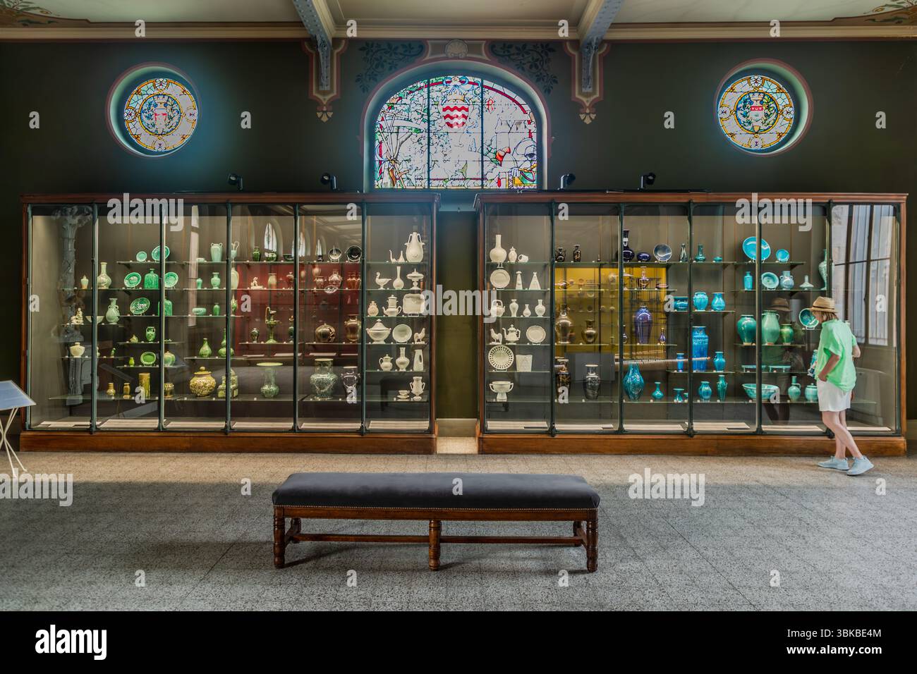 A display case with porcelain objects sorted by colour stands in the Musée National Adrien Dubouché on Place Winston Churchill in Limoges, Haute-Vienne, Nouvelle-Aquitaine, France. Two long glass cabinets hold vases, jugs, plates and figurines, arranged from white and celadon green on the left to dark, turquoise and blue glazes on the right. Journalist Angela Berg, wearing a straw hat, a light green blouse and white shorts, stands at the right-hand cabinet and looks at the objects. Above the cabinets are an arched stained-glass window with a coat of arms and two round stained-glass medallions. Stock Photo