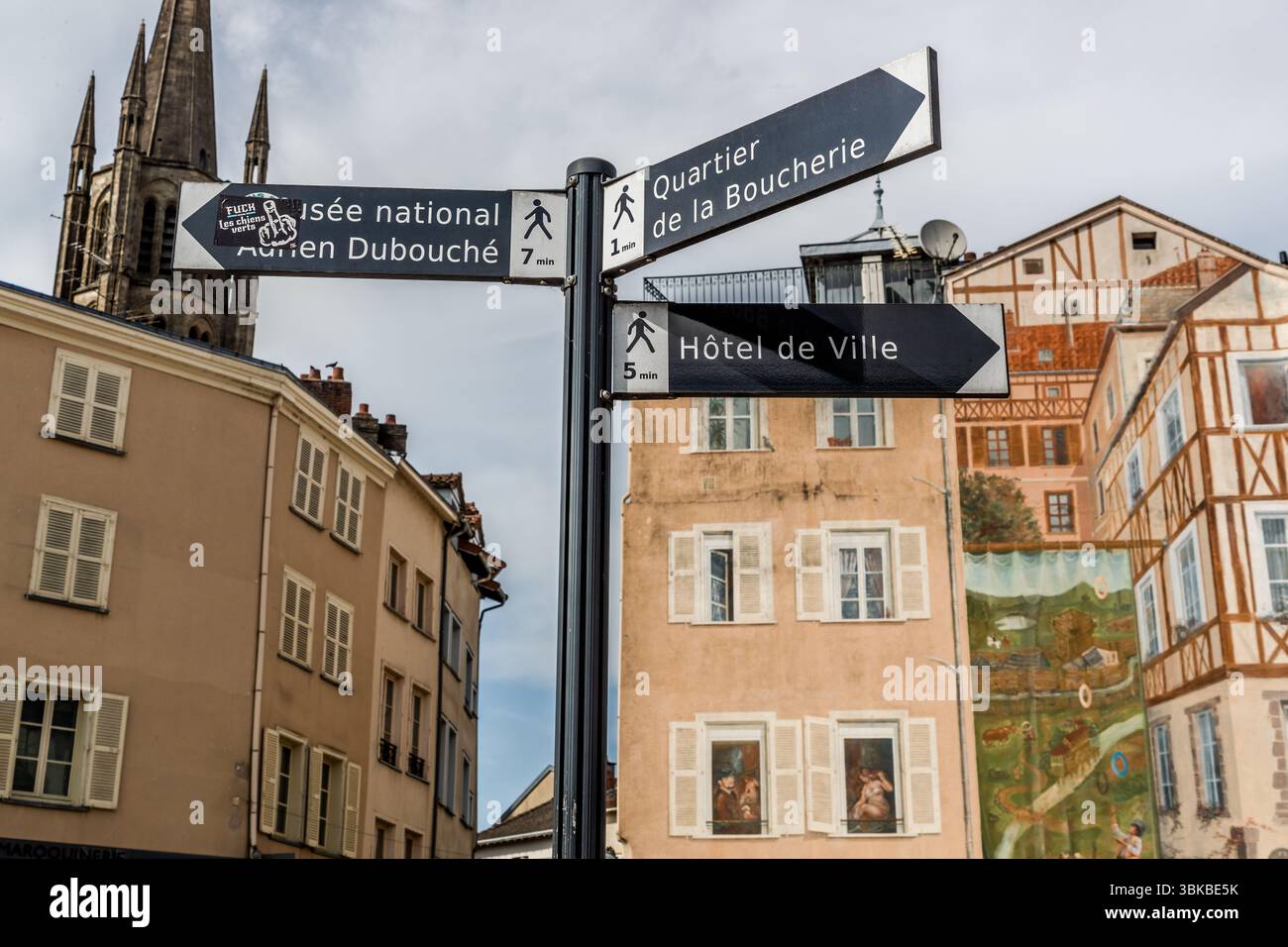 Signposts in the centre of Limoges, Haute-Vienne, Nouvelle-Aquitaine, France, on Rue Adrien Dubouché point to the city's sights. They show the way to the Musée National Adrien Dubouché (seven minutes on foot), the butchers' quarter (Quartier de la Boucherie, one minute) and the town hall (Hôtel de Ville, five minutes). A sticker covers part of the lettering for the museum. In the background, a mural shows motifs in the style of the old half-timbered houses. The tower of a Gothic church (possibly Saint-Michel-des-Lions) rises on the left. Stock Photo