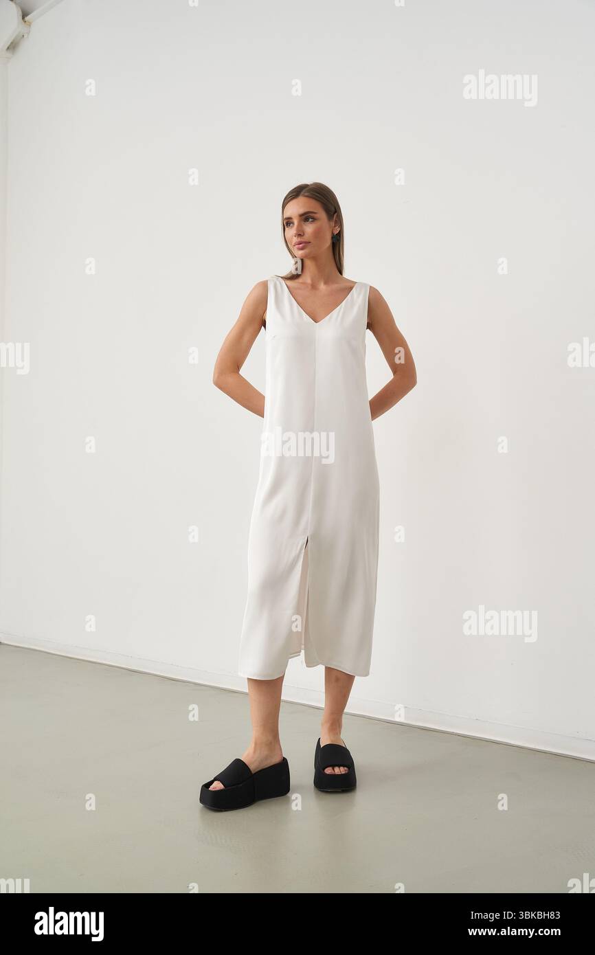 Woman in a sleeveless white dress and black platform shoes poses confidently. Her hands rest behind her back, and her gaze is forward. Minimalist Stock Photo