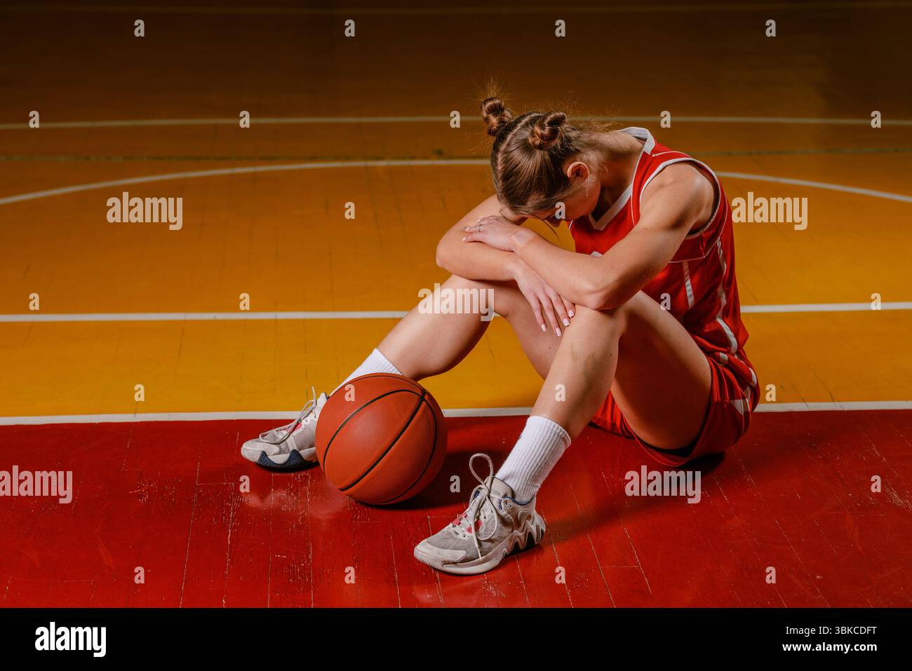 A dejected female basketball player sits on a court with her head down, resting on her knees, next to a basketball. Stock Photo