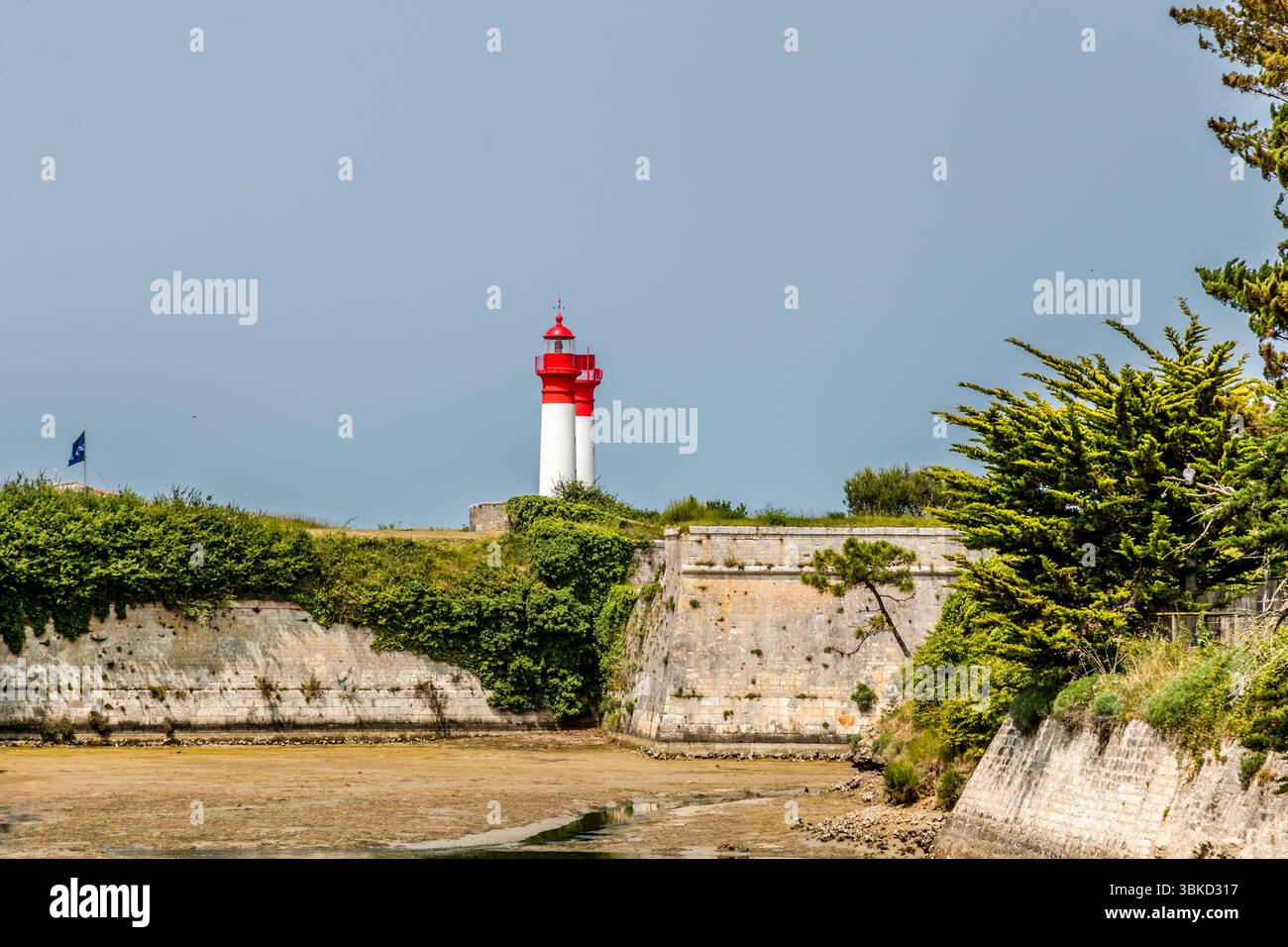 The fortification walls of Fort de la Rade on the island of Aix (Île d'Aix), Charente-Maritime, Nouvelle-Aquitaine, France, are overlooked by the island's two red and white lighthouses. In front of the walls lies a muddy channel, and a blue flag flies on the rampart on the left. Pines and bushes grow on the slope on the right. The lighthouses guide ships safely through the shallow and sometimes dangerous waters around the island, off the mouth of the Charente. Access to the Rochefort naval arsenal had to be well protected and controlled. For this reason, strategic light points were set up. The Stock Photo