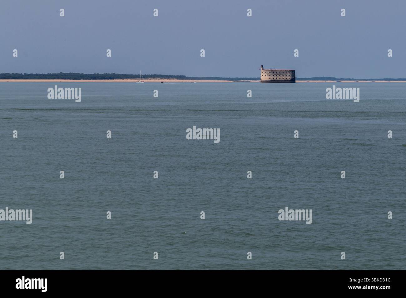 Fort Boyard stands on a sandbank in the sea between the island of Aix (Île d'Aix) and the Île d'Oléron, Charente-Maritime, Nouvelle-Aquitaine, France. The fort is clearly visible from the island's ferry landing. Behind it stretches a coast with a sandy beach and forest, and a sailing boat lies on the water to the left. Fort Boyard can only be visited by boat. It gained international fame through the adventure game show "Fort Boyard". The format was adapted in over 33 countries from 1990 onwards. It is considered the most successful French television export, with a huge international audience.  Stock Photo