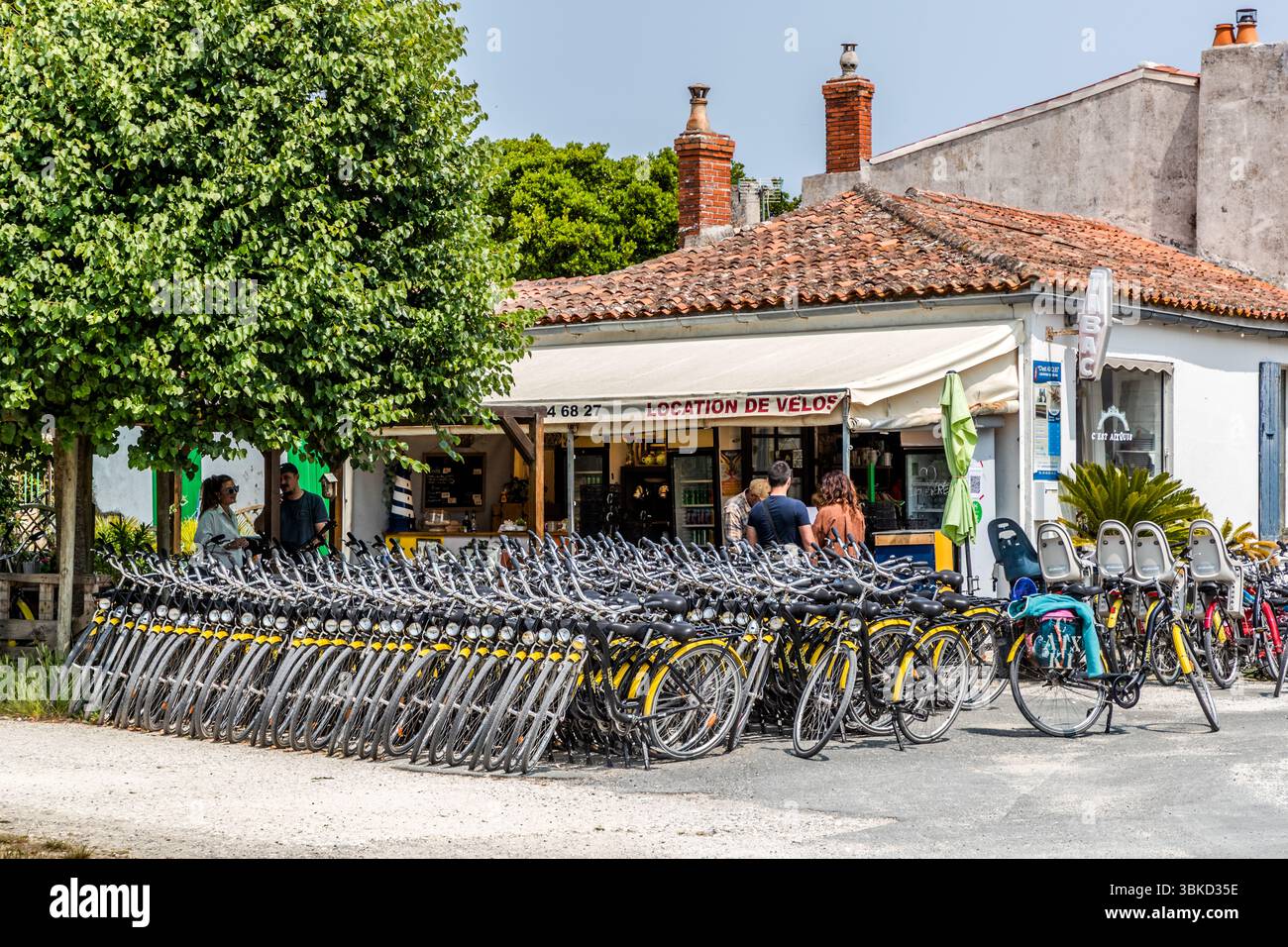 A bicycle rental shop with the sign "Location de vélos" stands on Place Austerlitz in Le Bourg on the island of Aix (Île d'Aix), Charente-Maritime, Nouvelle-Aquitaine, France. In English: "Bicycle rental". Long rows of yellow and grey rental bikes stand in front of the building with its tiled roof, some fitted with child seats. Customers wait at the counter under the awning. The island is car-free and, measuring 3 kilometres long and 600 metres wide, is perfect for cyclists and pedestrians. Stock Photo