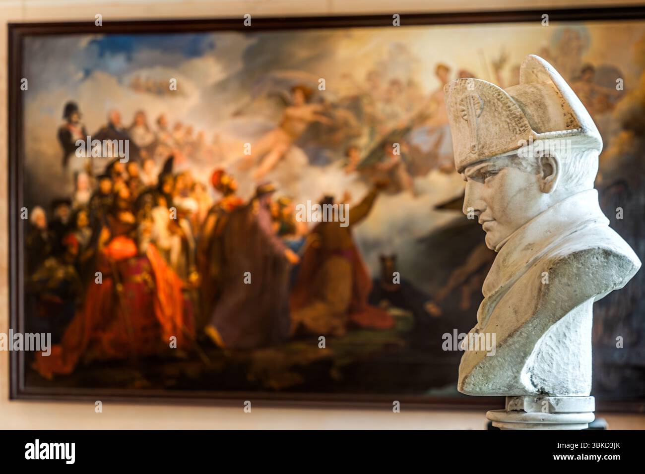 A bust of Napoleon Bonaparte stands in front of a large painting in the Napoleon Museum (Musée national napoléonien) on Rue Napoléon on the island of Aix (Île d'Aix), Charente-Maritime, Nouvelle-Aquitaine, France. The white bust shows the emperor in profile, wearing his bicorne hat. The painting behind it shows a crowd of figures in colourful robes beneath clouds with angels. The island played a special role in Napoleon's military strategy. After inspecting the island in 1808, Napoleon Bonaparte ordered the construction of massive fortifications to protect the nearby naval arsenal of Rochefort Stock Photo