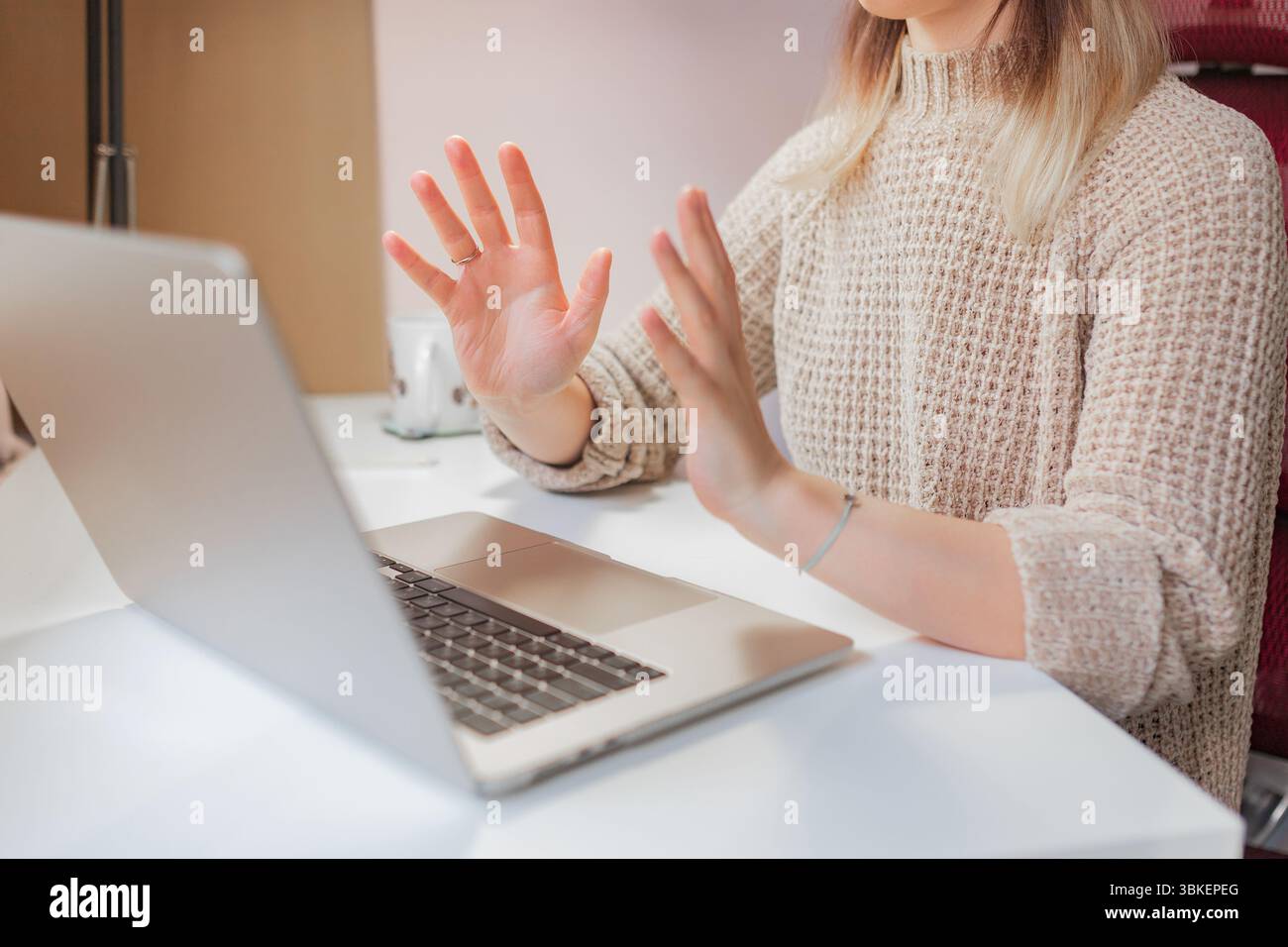 Woman holds up hands while communicating during remote meeting on laptop at home. Two open palms facing screen, woman speaks using expressive hand ges Stock Photo