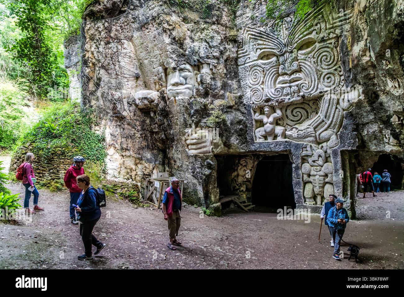 The Lapidiales sculptures in Port-d'Envaux, Nouvelle-Aquitaine, France, include a large face in the style of Māori carving with spiral patterns on a rock face (possibly a work by the New Zealand sculptor Paora Toi Te Rangiuaia). Below it are a female figure with raised arms, a lizard and a stylised figure next to a dark opening in the rock. On the left, a head protrudes from the rock above a large hand. Journalist Angela Berg (left, with a red backpack) holds a sheet of paper while visitors walk across the floor of the quarry. Various artists have carved the Lapidiales into the rock. The works Stock Photo