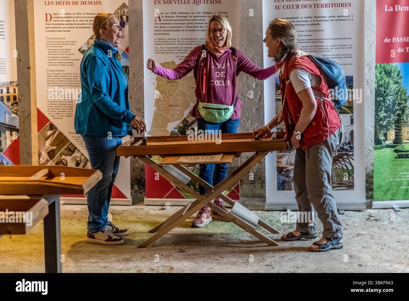 Journalist Angela Berg, in the centre, plays with two fellow travellers at a wooden games table in the collection of medieval games at La Roche Courbon Castle (Château de la Roche Courbon) on the Charente, Rue de la Belle au Bois Dormant, Saint-Porchaire, Nouvelle-Aquitaine, France. The three women are laughing. After a guided tour of the castle, the visit ends in the games collection. Guided tours of part of the family-owned and inhabited castle are available. Behind the women stand exhibition panels of the association La Demeure Historique, which has represented privately owned historic monu Stock Photo