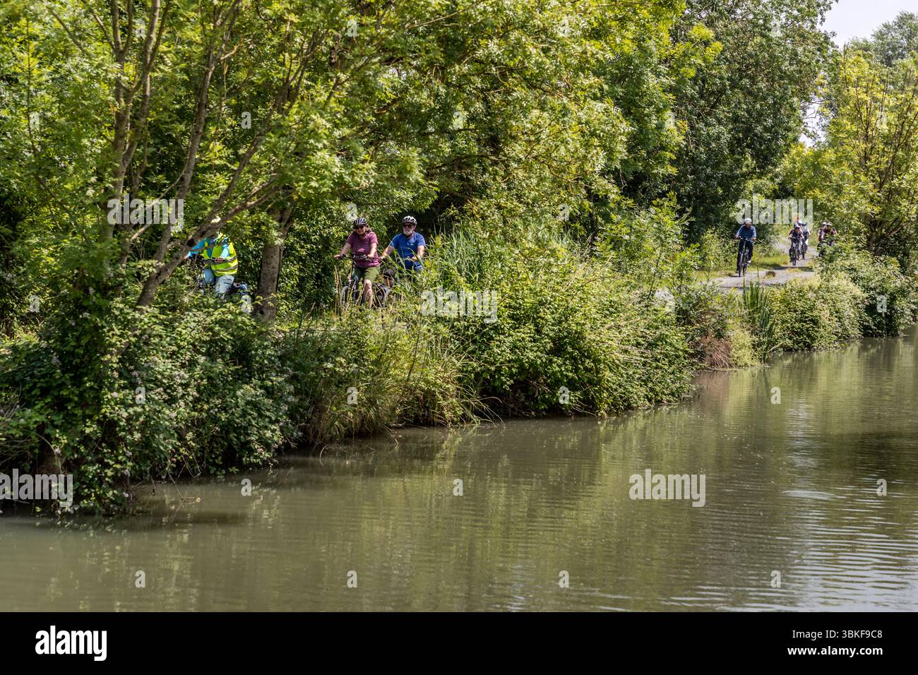 The FlowVélo long-distance cycle route (La Flow Vélo) runs for long stretches along the Charente River in the Nouvelle-Aquitaine region. Participants in the guided cycling tour by Die Landpartie ride along a bank path beside the water (possibly a canal parallel to the Charente), with tour guide Katell Nicoux in a high-visibility vest on the left. Trees and reeds line the bank at Rue de la Rivière, La Vallée, France. The route runs for around 400 kilometres from Sarlat-la-Canéda in the Périgord to the Île d'Aix. Stock Photo