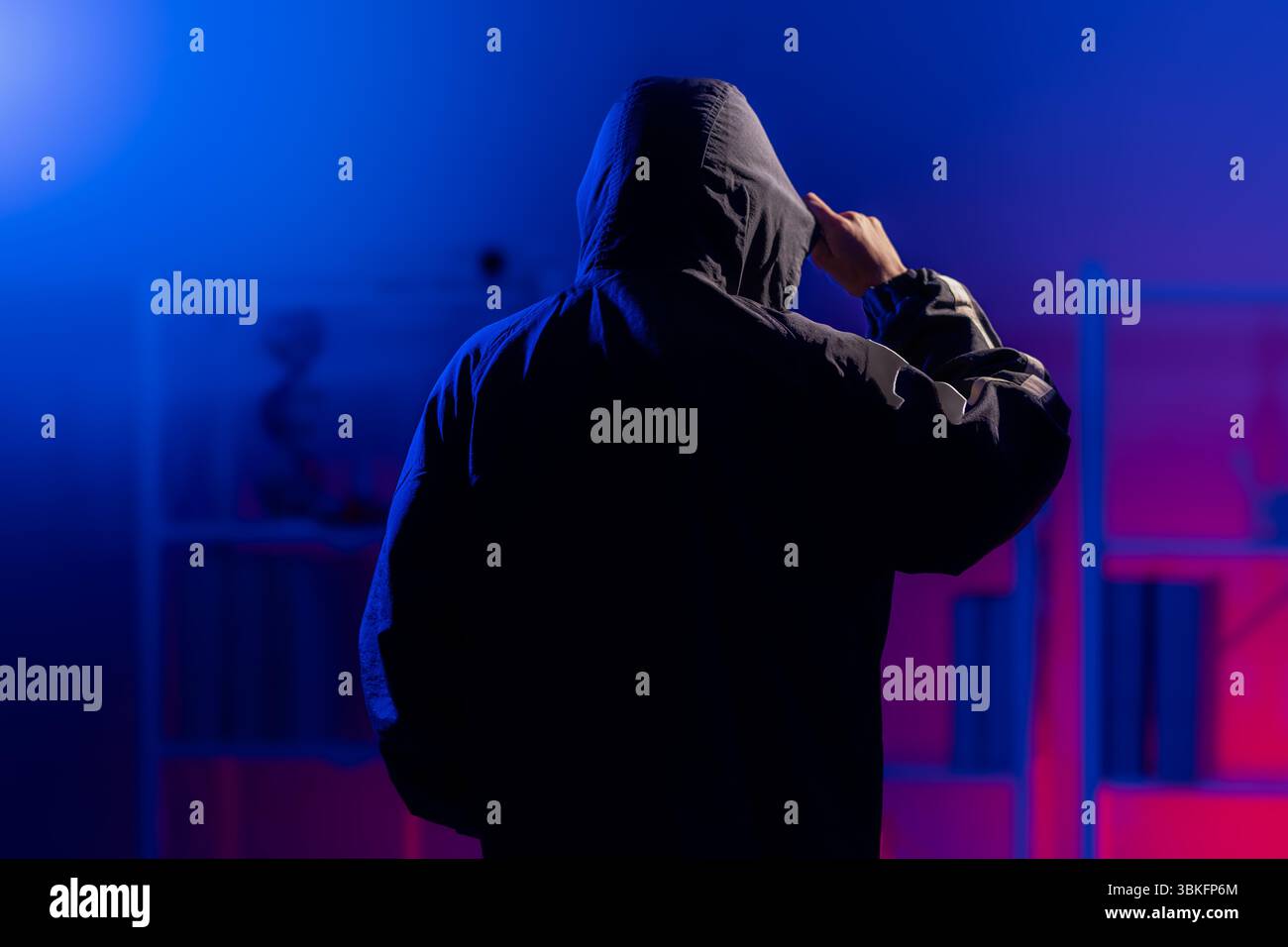 Back view of a man in a black hoodie standing in a dark neon-lit room, pulling down on her hoodie to cover his face, evoking themes of cybersecurity, Stock Photo